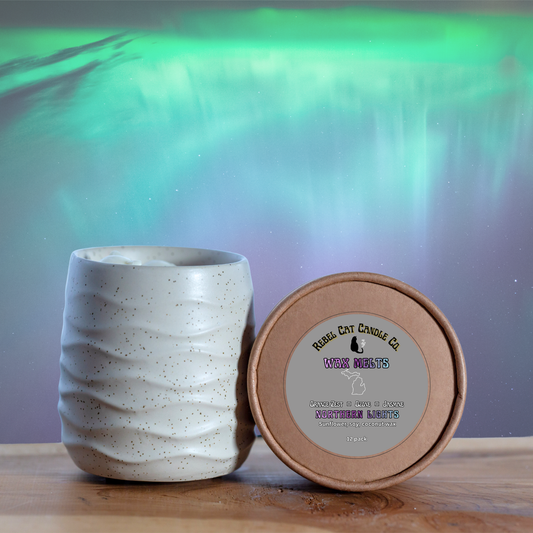 A ceramic candle tin with a wavy ribbed texture in a warm off-white glaze stands next to its round kraft paper lid. The lid features a circular label for 'Rebell Cat Candle Co.