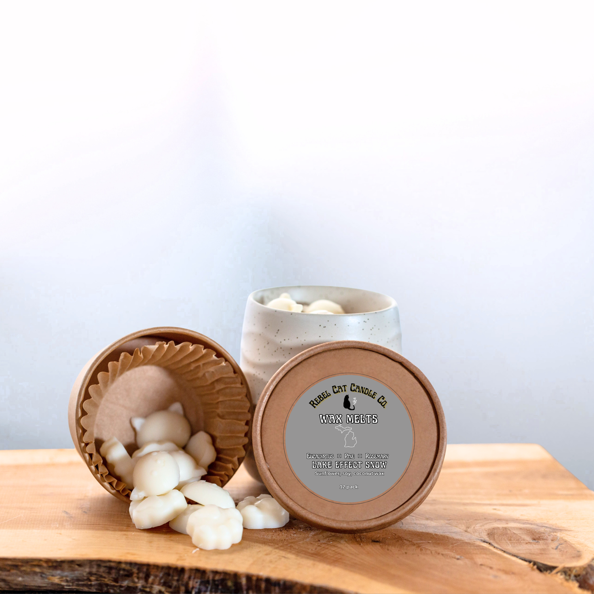 A ceramic wax melts jar  with a round wooden lid in front, showing white wax melts spilling out onto a wooden surface. The candle is labeled 'Real Cat Candles Co.