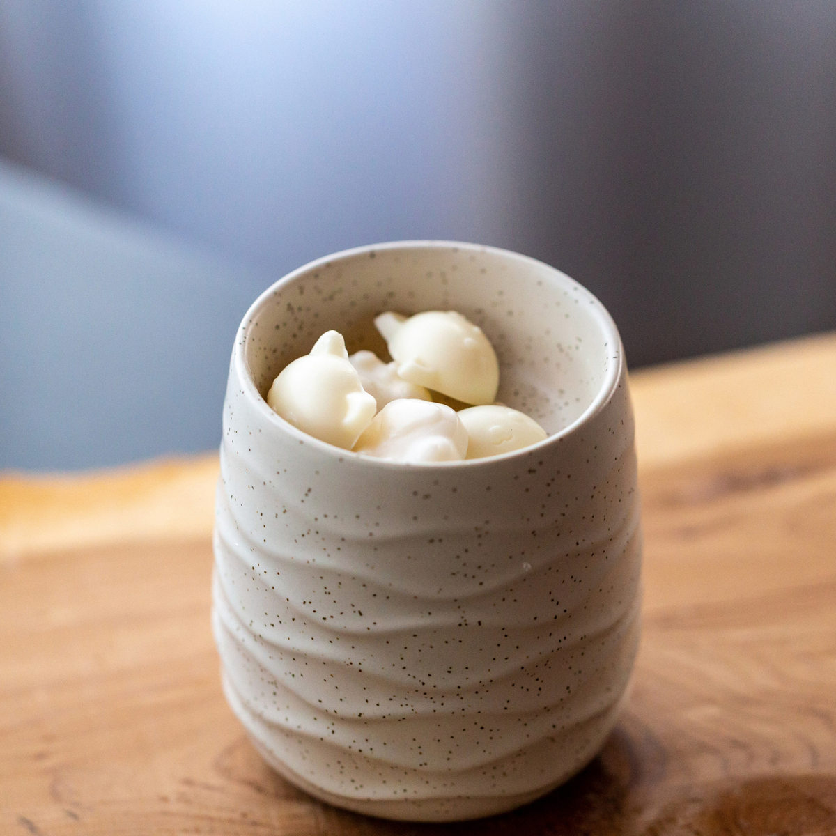 A small speckled ceramic cup filled with white cat shaped wax melts set on a wooden surface with a softly blurred background.