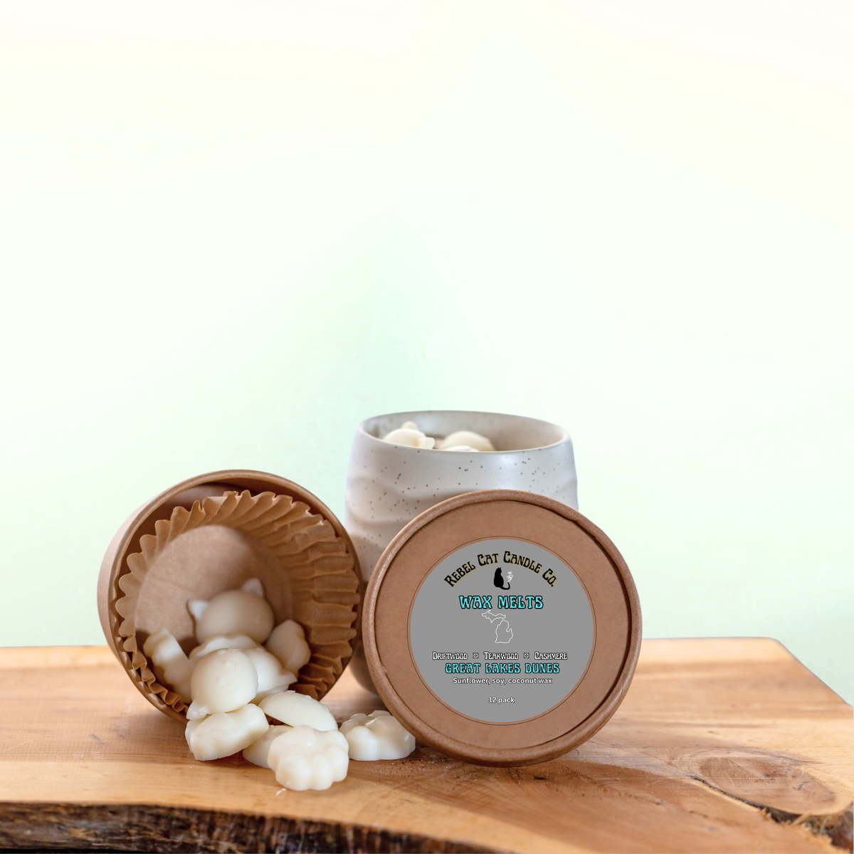 A ceramic jar of wax melts sits on a wooden surface with one lid leaning beside it and several white wax melts spilled out. The lid label reads 'Rebel Cat Candle Co.