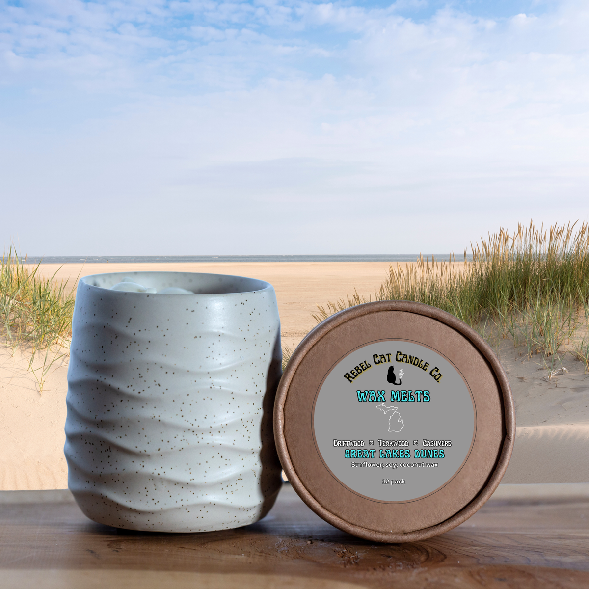 A ceramic wax melts pot with a textured, speckled light gray exterior sits next to its round brown cardboard lid on a wooden surface, with a beach landscape in the background.