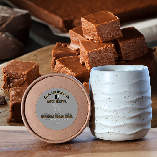 A ceramic candle jar with a speckled off-white exterior and a brown lid sits in front of a stack of chocolate brown fudge bars on a wooden surface. The lid label reads 'Rebel Cat Candle Co.