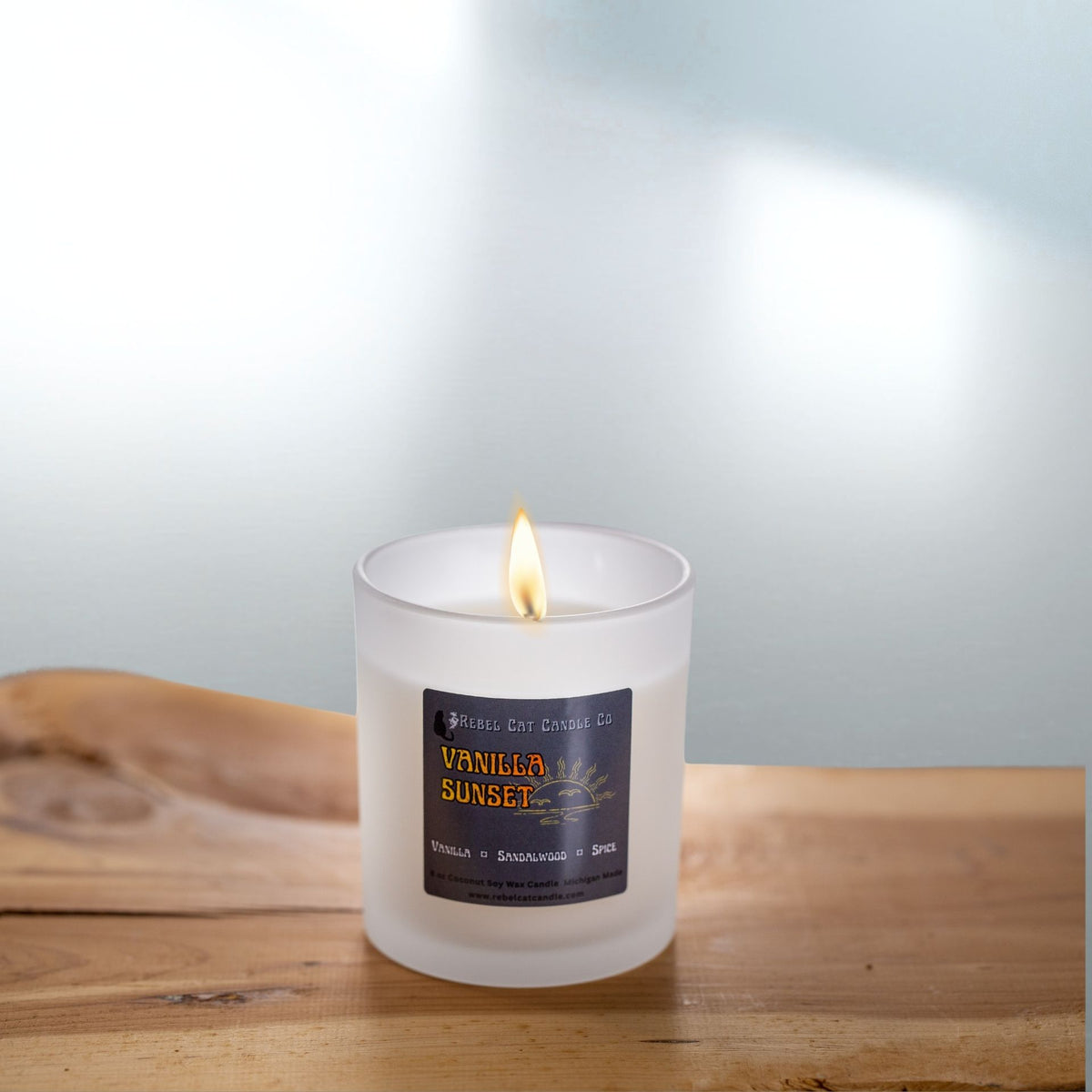 Frosted white candle with label by Rebel Cat Candle titled Vanilla Sunset on a table with soft background