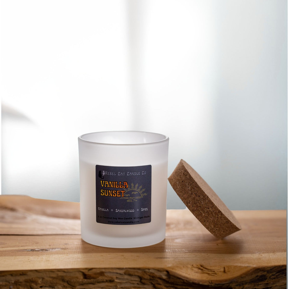 Frosted glass candle jar with cork lid leaning to the side on a wood table with soft white curtains in the background