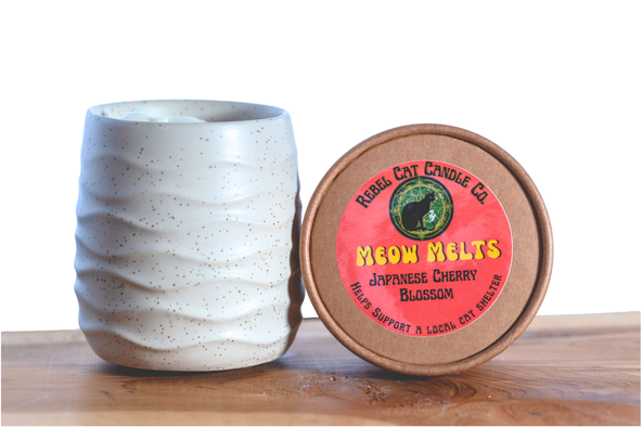 A ceramic warmer  jar with a wavy, speckled cream and brown glaze sits next to a round lid. The lid features a colorful label with a coral pink background, yellow text reading 'MEOW MELTS', and a green cat silhouette logo.