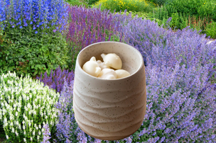 A small beige textured ceramic cup holding a few white wax melts set against a blurred garden background of purple flowers and green foliage.