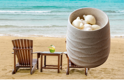 A large speckled beige ceramic mug filled with white wax melts placed on a sandy beach next to two wooden lounge chairs and a small table with a green drink, with a lake in the background.
