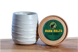 A small textured ceramic mug in a light speckled gray-white glaze sits next to a round lid, which features a green label reading 'MEOW MELTS' on a wooden surface against a white background.