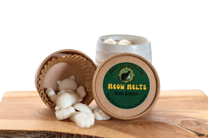 A jar of Meow Melts melts with one lid removed and tipped over to show the white melts inside, placed on a wooden board against a white background.